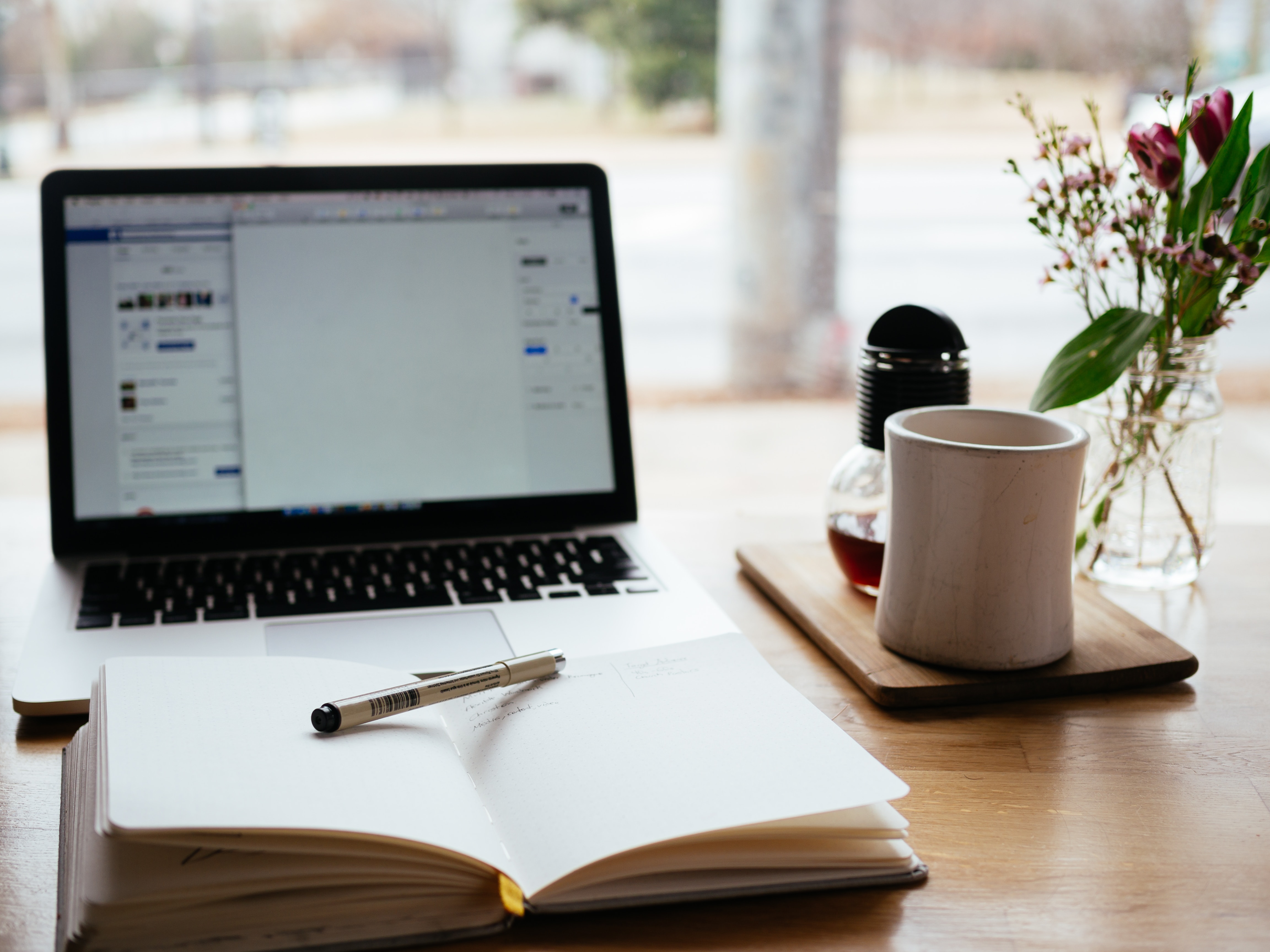 Um computador com um caderno e caneta a sua frente, ao lado uma caneca com chá e um vaso de flores